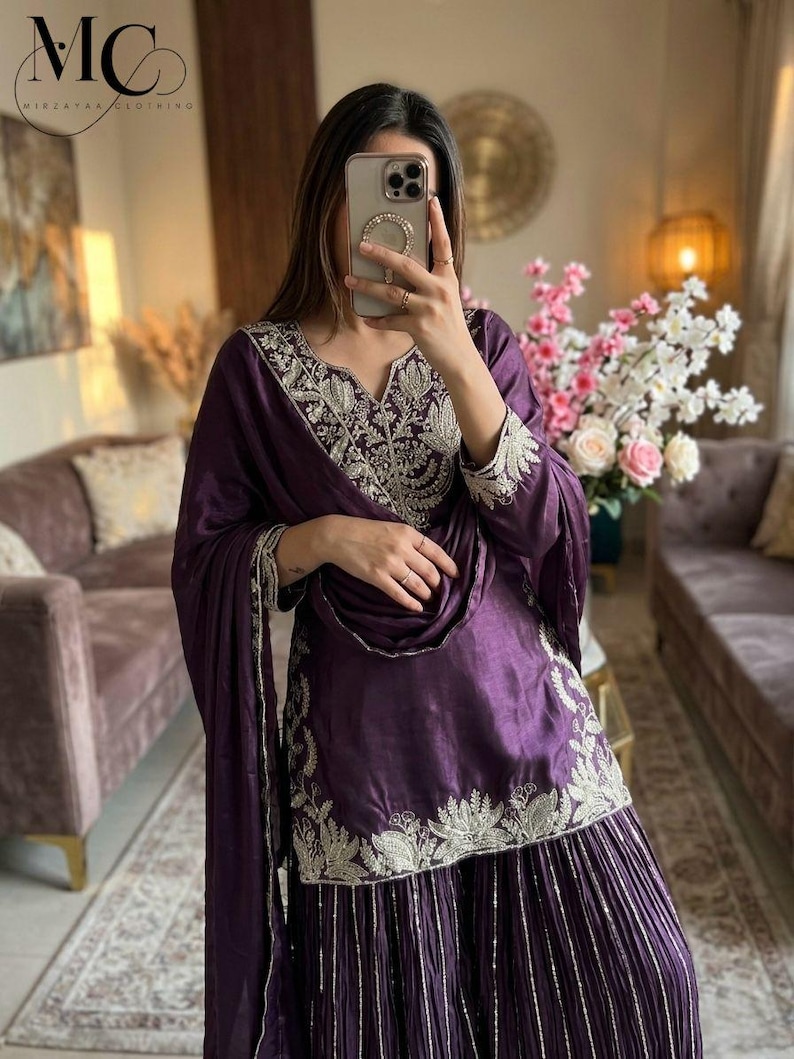 Może przedstawiać: Głęboki fioletowy, haftowany zestaw, składający się z tuniki, spodni z szerokimi nogawkami i pasującego szalika. Str&oacute;j ma skomplikowane srebrne hafty wok&oacute;ł dekoltu, rękaw&oacute;w i dołu. Widoczne jest logo marki "MC MIRZAYAA CLOTHING".