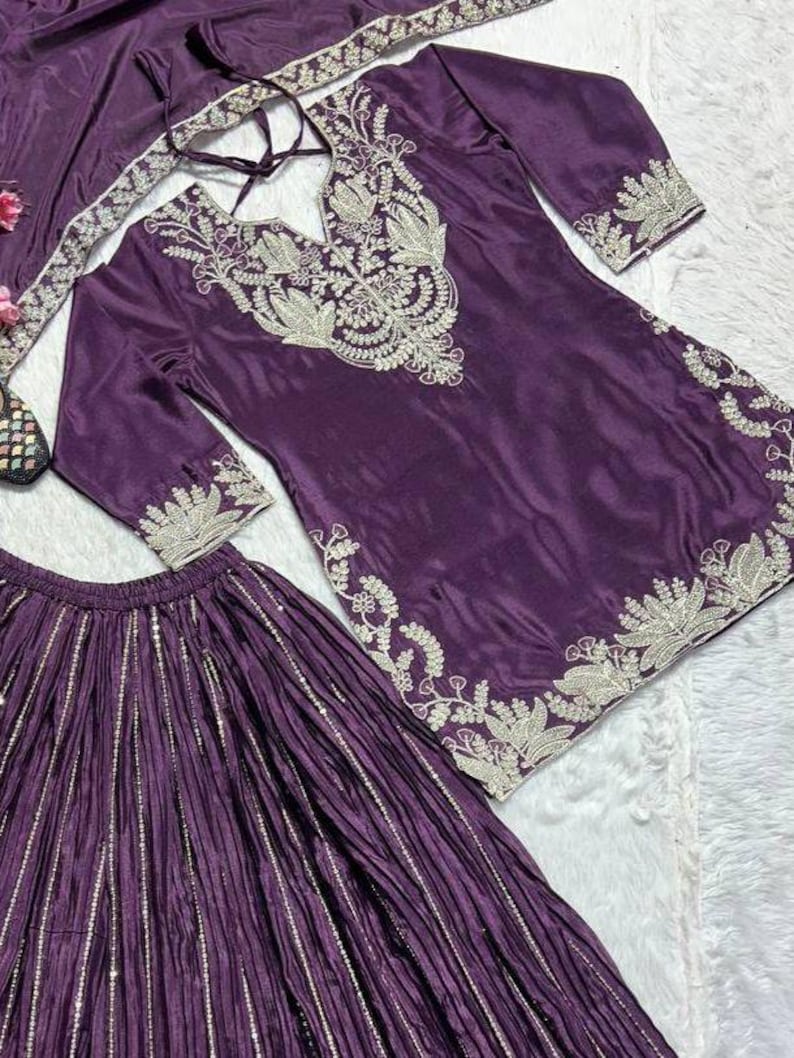 Może przedstawiać: Śliwkowy, trzyczęściowy str&oacute;j składający się z tuniki, sp&oacute;dnicy i szalika. Tunika ma skomplikowane srebrne hafty wok&oacute;ł dekoltu, rękaw&oacute;w i dołu. Sp&oacute;dnica jest plisowana z pionowymi srebrnymi paskami. Szalik r&oacute;wnież ma srebrne hafty.