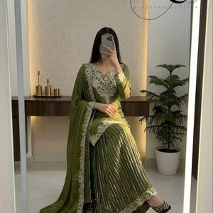 Peut inclure: Ensemble vert olive compos&eacute; d'un haut &agrave; manches longues avec broderies d&eacute;taill&eacute;es et d'une jupe pliss&eacute;e. L'ensemble comprend un ch&acirc;le assorti avec des bordures brod&eacute;es. L'image montre &eacute;galement des chaussures plates &agrave; motifs noirs et blancs. Le logo de la marque "MC Mirzayaa Clothing" est visible.