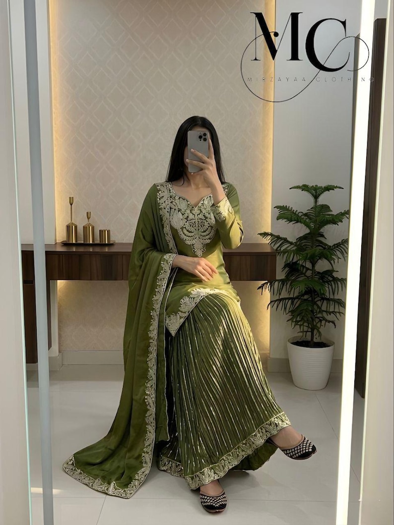 Peut inclure: Ensemble vert olive compos&eacute; d'un haut &agrave; manches longues avec broderies d&eacute;taill&eacute;es et d'une jupe pliss&eacute;e. L'ensemble comprend un ch&acirc;le assorti avec des bordures brod&eacute;es. L'image montre &eacute;galement des chaussures plates &agrave; motifs noirs et blancs. Le logo de la marque "MC Mirzayaa Clothing" est visible.