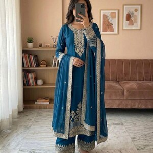 May include: A teal embroidered salwar kameez outfit. The set features a long-sleeved tunic, wide-leg trousers, and a matching dupatta. The intricate gold embroidery decorates the neckline, sleeves, and hemline, with matching gold flats.