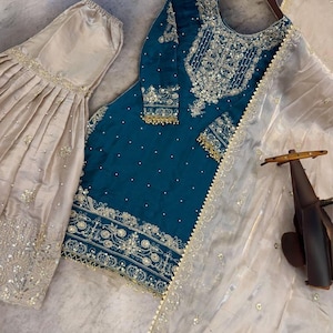 Puede incluir: Un elegante conjunto bordado de color verde azulado con una bufanda transparente a juego y una prenda plisada beige. El conjunto verde azulado presenta intrincados bordados plateados y dorados. La bufanda transparente tiene un ribete dorado.