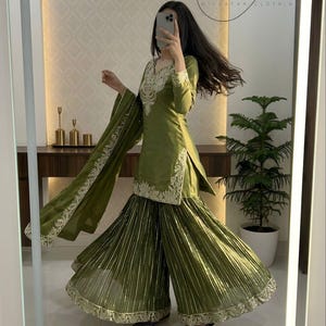 Peut inclure: Ensemble sharara vert olive avec broderies d&eacute;taill&eacute;es sur l'encolure et les manches. Le pantalon large pr&eacute;sente des plis verticaux et une bordure dor&eacute;e. La tenue comprend un dupatta assorti avec une bordure d&eacute;taill&eacute;e. Le texte "MC" et "MISEAVAA CLOTHING" est visible.