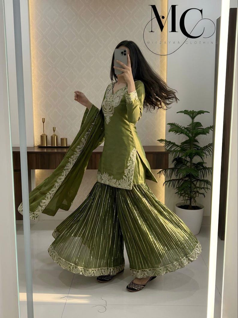 Peut inclure: Ensemble sharara vert olive avec broderies d&eacute;taill&eacute;es sur l'encolure et les manches. Le pantalon large pr&eacute;sente des plis verticaux et une bordure dor&eacute;e. La tenue comprend un dupatta assorti avec une bordure d&eacute;taill&eacute;e. Le texte "MC" et "MISEAVAA CLOTHING" est visible.