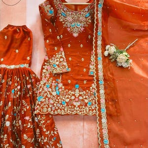 May include: Rust-colored embroidered salwar kameez set. The ensemble features intricate floral embroidery in shades of blue, pink, and white. The set includes a long-sleeved top, a flared skirt, and a matching dupatta.