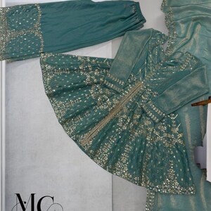Pu&ograve; includere: Completo formale color teal con intricati abbellimenti dorati. L'ensemble comprende una tunica a maniche lunghe, pantaloni abbinati e una sciarpa drappeggiata. La tunica presenta un motivo dettagliato di paillettes e ricami dorati. Il logo del marchio "MC MIRZAYAA CLOTHING" &egrave; visibile.