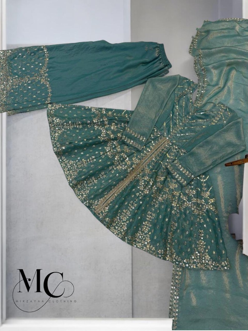 Pu&ograve; includere: Completo formale color teal con intricati abbellimenti dorati. L'ensemble comprende una tunica a maniche lunghe, pantaloni abbinati e una sciarpa drappeggiata. La tunica presenta un motivo dettagliato di paillettes e ricami dorati. Il logo del marchio "MC MIRZAYAA CLOTHING" &egrave; visibile.