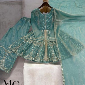 Pu&ograve; includere: Un completo formale ricamato color verde acqua, composto da un top a maniche lunghe, pantaloni e una sciarpa abbinata. Il top presenta intricati abbellimenti argentati e una silhouette svasata. L'outfit &egrave; esposto su un'appendiabiti su sfondo neutro. Il logo del marchio "MC MIRZAYAA CLOTHING" &egrave; visibile.