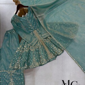 Pu&ograve; includere: Un completo formale color verde acqua, impreziosito, composto da un top a maniche lunghe, pantaloni e una sciarpa abbinata. Il top presenta ricami argentati e dettagli di paillettes. L'outfit &egrave; esposto su una gruccia di legno. Il logo "MC MIRZAYAA CLOTHING" &egrave; visibile.