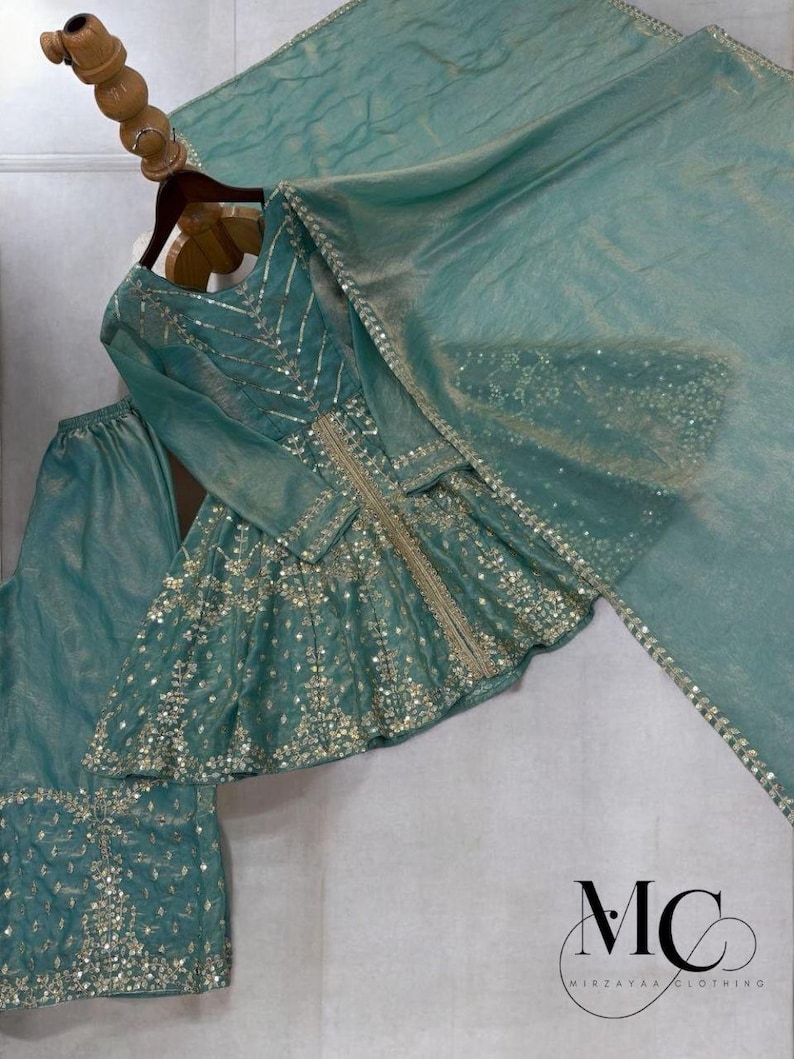 Pu&ograve; includere: Un completo formale color verde acqua, impreziosito, composto da un top a maniche lunghe, pantaloni e una sciarpa abbinata. Il top presenta ricami argentati e dettagli di paillettes. L'outfit &egrave; esposto su una gruccia di legno. Il logo "MC MIRZAYAA CLOTHING" &egrave; visibile.