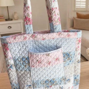 May include: Quilted tote bag with a patchwork design. The bag features a combination of pink, blue, and white floral and polka dot patterns. It has a front pocket and sturdy handles, perfect for carrying everyday essentials.