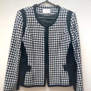 May include: A monochrome houndstooth blazer with black detailing and two front pockets. The jacket is suspended from a black hanger. The blazer features a round neckline and long sleeves.