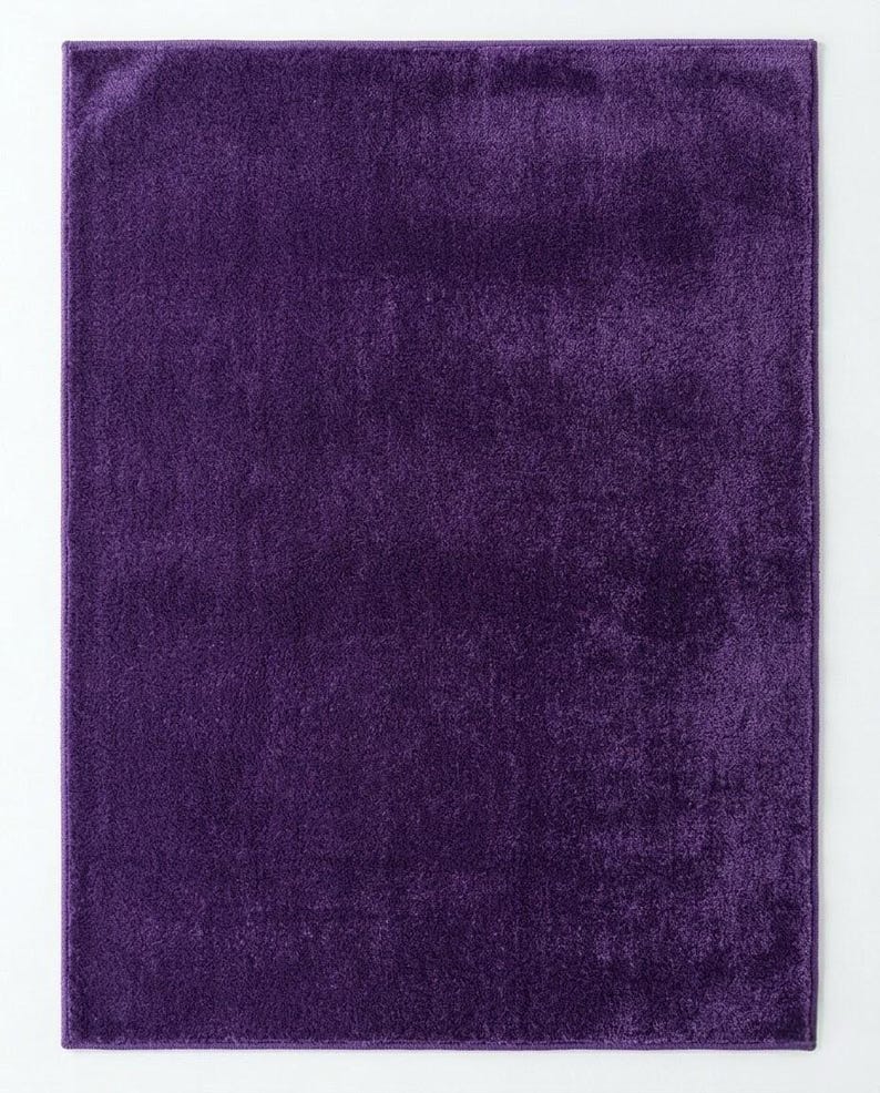 May include: A rectangular, deep purple rug with a soft, velvety texture. The rug has a solid colour and is viewed from above, showcasing its smooth surface and rich hue. The rug is a solid colour, suitable for various interior settings.