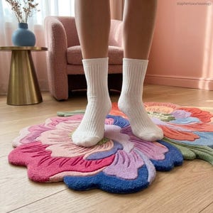 K&ouml;nnte beinhalten: Ein mehrfarbiger, bl&uuml;tenf&ouml;rmiger Teppich in Rosa-, Blau- und Oranget&ouml;nen. Der Teppich besteht aus einem weichen, strukturierten Material. Wei&szlig;e Socken stehen auf dem Teppich. Der Teppich ist ein dekorativer Bodenbelag.
