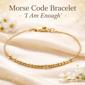 Könnte beinhalten: Goldfarbenes Morsecode-Armband mit der Botschaft 'I Am Enough'. Das Armband hat eine feine Kette und goldene Perlen und Stäbe, die den Morsecode bilden. Es wird auf einem cremefarbenen Satinhintergrund mit weißen Blumen präsentiert. Ein stilvolles Schmuckstück.