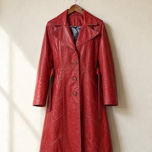 May include: A long, red leather trench coat featuring a wide collar, a button-front closure with three visible brown buttons, long sleeves, and a matching tie belt at the waist. The coat has diagonal seam detailing and a dark patterned lining.