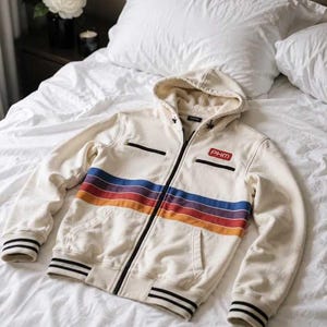 Op de afbeelding: Een gebroken witte hoodie met rits en een horizontaal strepenpatroon in blauw, paars en oranje over de borst. Het kledingstuk is voorzien van een rode rechthoekige patch met de tekst PHM op de linkerborst, zwarte ritsdetails en zwart gestreepte boorden.