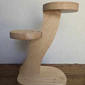 May include: A wooden display stand with a natural finish. It features a curved support with two circular platforms and a rectangular base. The stand is made of light-colored wood and has a smooth, polished appearance.