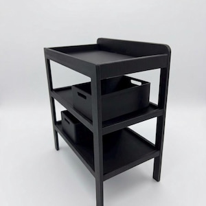 May include: A black wooden changing table featuring a raised top edge, two lower open shelves, and two rectangular black storage bins with cutout handles placed on the middle and bottom shelves. The structure has four straight legs and a frame.