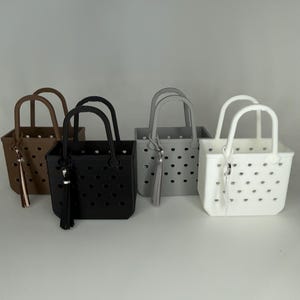 May include: Four small tote bags in brown, black, gray, and white. Each bag has a rectangular shape with a handle and a tassel. The bags have a perforated design and are displayed on a white surface.