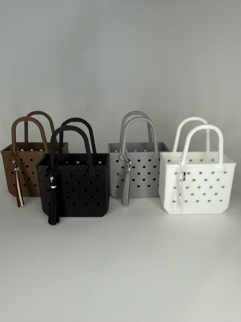 May include: Four small tote bags in brown, black, gray, and white. Each bag has a rectangular shape with a handle and a tassel. The bags have a perforated design and are displayed on a white surface.