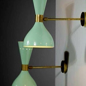 Puede incluir: Dos apliques de pared verde menta con forma de reloj de arena, acentuados con metal dorado. Los apliques tienen un diseño retro, con pequeños puntos blancos alrededor de la parte superior e inferior. Los apliques están montados en una pared blanca.