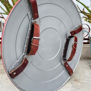 K&ouml;nnte beinhalten: Ein silberfarbener, runder Schild mit rotem Rand. Er hat braune Lederriemen und Metallbeschl&auml;ge. Das Design erinnert an einen Superheldenschild, mit einem Durchmesser von etwa 60 cm.