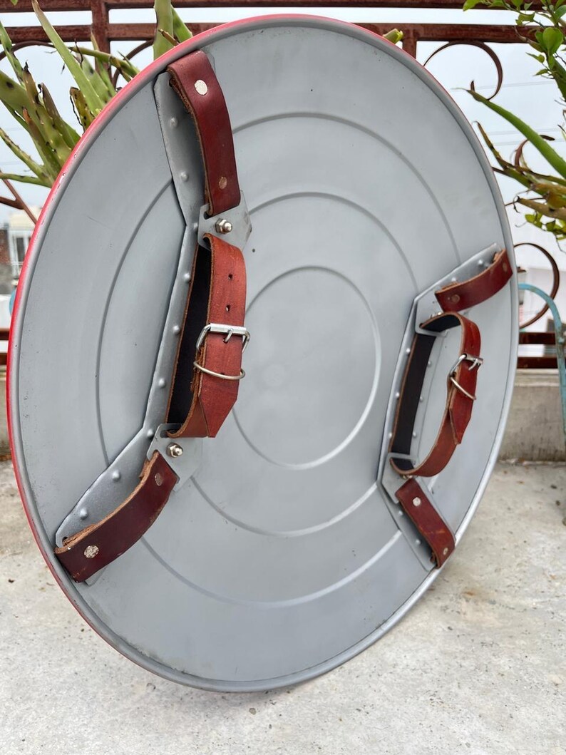 K&ouml;nnte beinhalten: Ein silberfarbener, runder Schild mit rotem Rand. Er hat braune Lederriemen und Metallbeschl&auml;ge. Das Design erinnert an einen Superheldenschild, mit einem Durchmesser von etwa 60 cm.