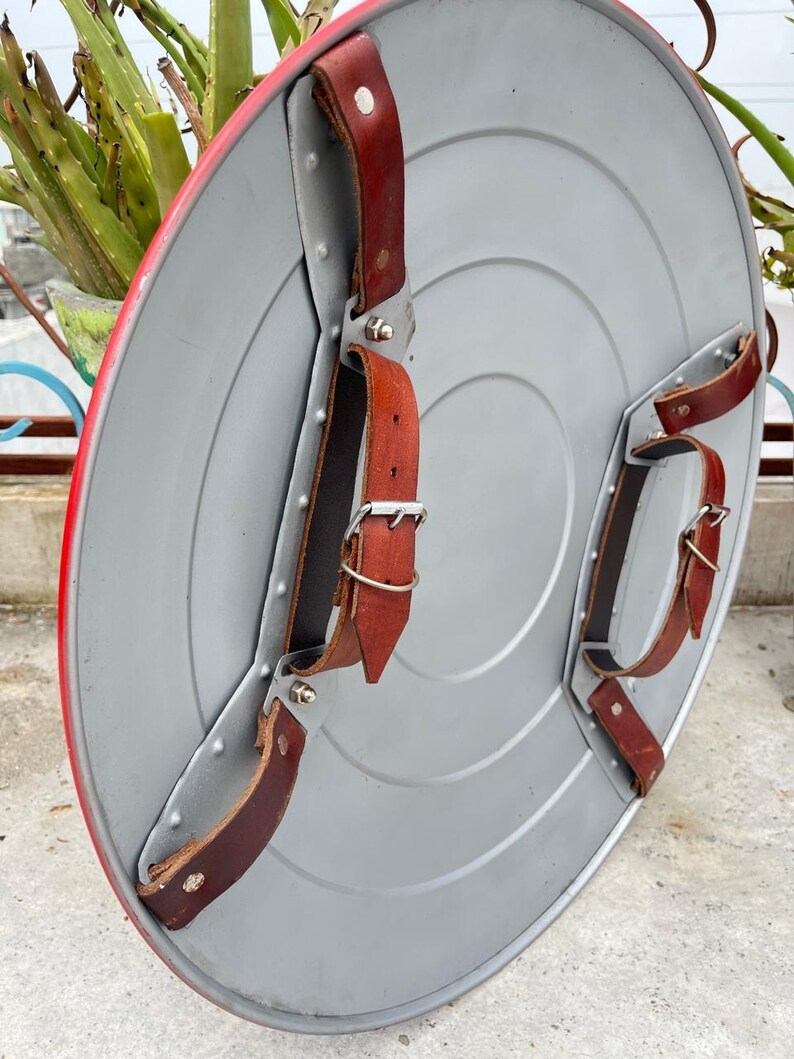 K&ouml;nnte beinhalten: Ein silberfarbener, kreisf&ouml;rmiger Schild mit rotem Rand. Der Schild hat konzentrische Kreise und ist mit braunen Lederriemen und Metallbeschl&auml;gen zur Befestigung am Arm ausgestattet. Ideal f&uuml;r Cosplay oder zur Pr&auml;sentation.