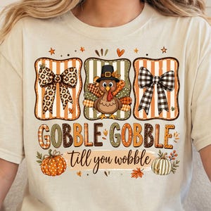 May include: A cream-colored short-sleeve t-shirt featuring a graphic print of a turkey wearing a pilgrim hat between two bows, one leopard print and one black and white plaid. The text reads GOBBLE GOBBLE till you wobble with pumpkins.