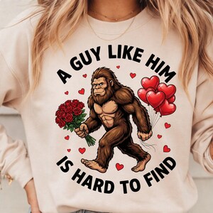 Puede incluir: Sudadera beige con un diseño de Bigfoot sosteniendo rosas rojas y globos con forma de corazón. El texto dice "A Guy Like Him Is Hard To Find", con detalles de corazones rojos. Un diseño divertido y romántico.