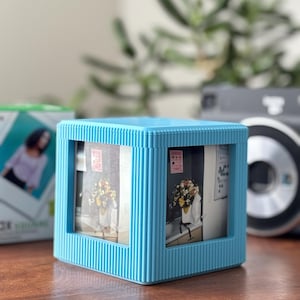 May include: A light blue cube-shaped photo frame with vertical ribbed detailing. The frame holds three visible photos of a floral arrangement. A box with a photo of a person is visible in the background, along with a camera.
