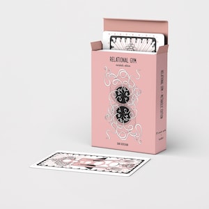 Pode incluir: Uma caixa de cartas rosa com a inscri&ccedil;&atilde;o "RELATIONAL GYM" e um baralho de cartas dentro. Uma carta est&aacute; colocada na frente. A caixa apresenta um design &uacute;nico com dois c&iacute;rculos pretos e um desenho linear branco. As cartas t&ecirc;m um design a preto e branco.