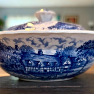 May include: A white ceramic serving bowl with a lid, featuring a blue transferware pattern depicting a detailed countryside scene with a timber-framed house, trees, and a cloudy sky. The bowl has a scalloped rim and a matching lid with a knob.