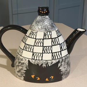 May include: A ceramic teapot featuring a black and white checkered pattern on the upper body and a speckled gray finish on the lower section. A black cat face with orange eyes is depicted on the front, and a small black cat figure sits on the lid.
