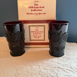 May include: Two dark red Avon 1876 Cape Cod Collection tall beverage glasses with decorative patterns. The glasses are displayed in front of their original box, which reads "The 1876 Cape Cod Collection Tall Beverage Glass Set (set of 2)".