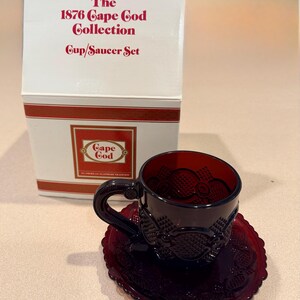 May include: A ruby red cup and saucer set from the 1876 Cape Cod Collection by Avon. The cup and saucer are made of glass and feature a textured design. The set is displayed in its original box.
