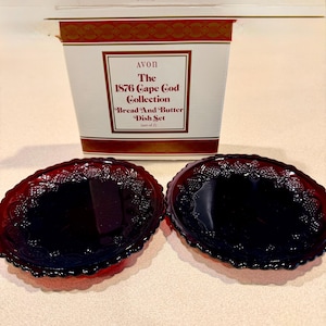 May include: A set of two ruby red glass bread and butter dishes from the Avon 1876 Cape Cod Collection. The dishes are oval-shaped with a decorative rim. The box is white with red and gold accents and features the collection name.