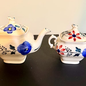 May include: A cream-colored ceramic teapot and sugar bowl set. The teapot has a curved spout and handle, and the sugar bowl has two handles. Both pieces are decorated with blue, red, and green floral designs.