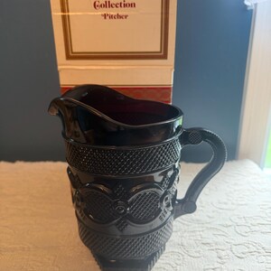May include: A deep red glass pitcher from the Avon The 1876 Cape Cod Collection, featuring a textured diamond pattern and a sturdy handle. The pitcher is displayed in front of its original white and red cardboard box with the printed text.