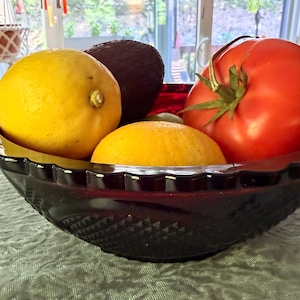 May include: A deep red glass bowl with a scalloped rim and a diamond-patterned exterior texture. The bowl is filled with a bright yellow lemon, a dark purple avocado, a smaller yellow citrus fruit, and a large, round, vibrant red tomato.