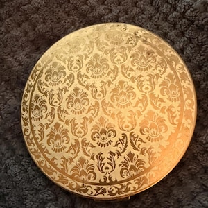 May include: A round, gold-toned compact with an ornate floral pattern. The compact's surface is covered in a repeating design of stylized flowers and leaves. The edges of the compact are also decorated with a similar pattern.