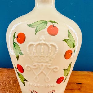 May include: Cream-colored ceramic bottle with a crown emblem and the dates 1898-1948. The bottle is decorated with red cherries and green leaves. The top of the bottle has a blue rim. The bottle is sitting on a wooden surface.