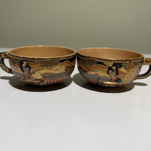May include: Two ceramic teacups featuring a dark exterior with gold trim and a hand-painted scene of a figure in traditional attire amidst a landscape. The interior of each cup is a light beige color, and each has a small, ornate handle on the side.