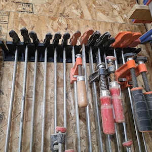 May include: A selection of woodworking clamps, including bar clamps and C-clamps, fixed to a wooden wall. The clamps are in various sizes and colours, with black, orange, and wooden handles. The image shows a workshop or garage setting.