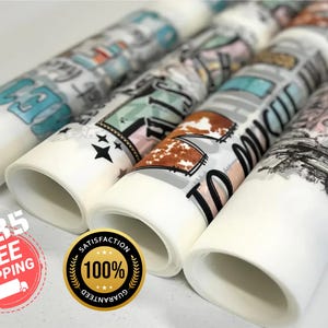 May include: Rolled decorative paper with various designs, including text and graphics. The designs feature a mix of colors, including teal, brown, and black. The paper rolls are white with the designs printed on them. The image shows several rolls.