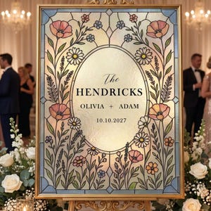 May include: A rectangular wedding sign in a gold frame featuring a stained glass design with pink and yellow flowers, green stems, and blue accents. The center oval contains the text: The HENDRICKS, OLIVIA + ADAM, and the date 10.10.2027.