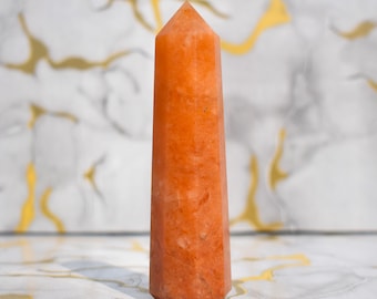 May include: A tall, orange crystal obelisk with a pointed top. The crystal has a smooth, polished surface and a warm, earthy tone. The obelisk is standing upright against a marble background with gold accents. This decorative item is a unique home decor piece.
