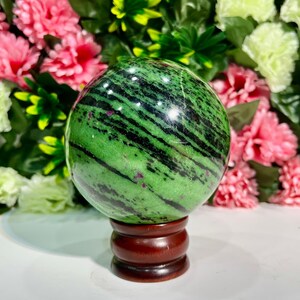 Può includere: Una sfera di zoisite verde lucidata con inclusioni nere e rosse, esposta su un supporto di legno marrone scuro. La superficie della sfera presenta un motivo a strisce. Lo sfondo presenta fiori rosa e verdi.