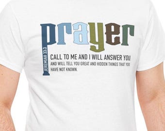 Puede incluir: Camiseta blanca con la palabra "prayer" en letras de bloque azules, verdes y marrones. La camiseta también presenta el texto "Call to me and I will answer you" y "Jeremiah 33:3".